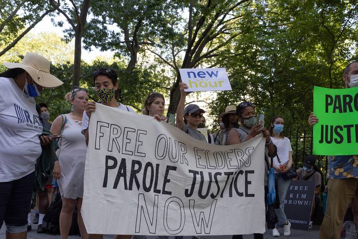 A handful of protestors hold a big white banner with the words "free our elders, parole justice now."